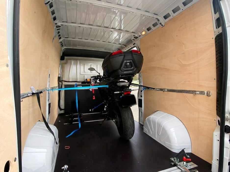 Intérieur de fourgon aménagé avec lambris bois clair et sol en caoutchouc noir, transportant une moto noire équipée d'un top-case