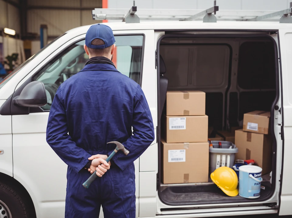 Technicien en combinaison bleue saluant depuis la portière ouverte d'un véhicule utilitaire blanc, tenant un rouleau de câble électrique ou de tuyauterie flexible